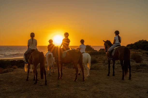 Plusieurs personnes à cheval sur une plage au coucher du soleil, la lumière chaude créant des silhouettes paisibles.