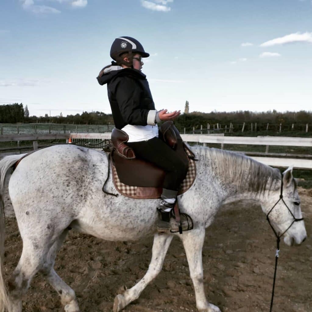 Jeune cavalier en casque sur un cheval gris pommelé en extérieur, mains ouvertes. Le cheval avance tranquillement tête baissée.
