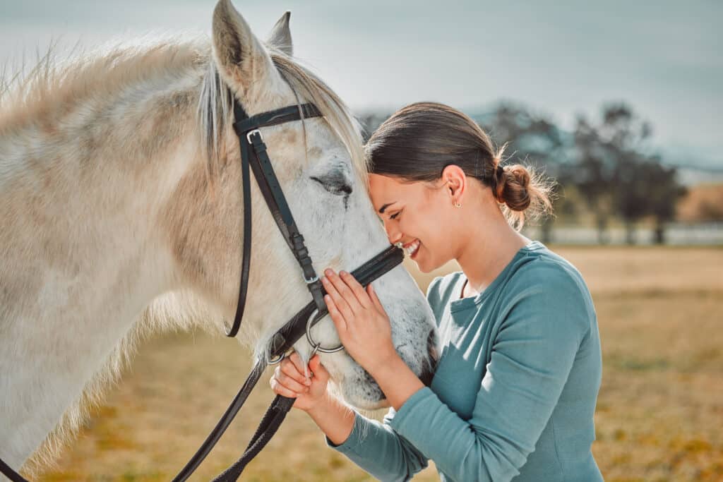 Femme souriante, la tête contre un cheval blanc au licol noir. Elle caresse son museau, exprimant une profonde affection en extérieur.