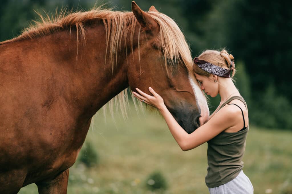 Jeune femme blonde caresse tendrement un cheval marron à crinière claire, fronts unis. Un instant de connexion paisible en nature.